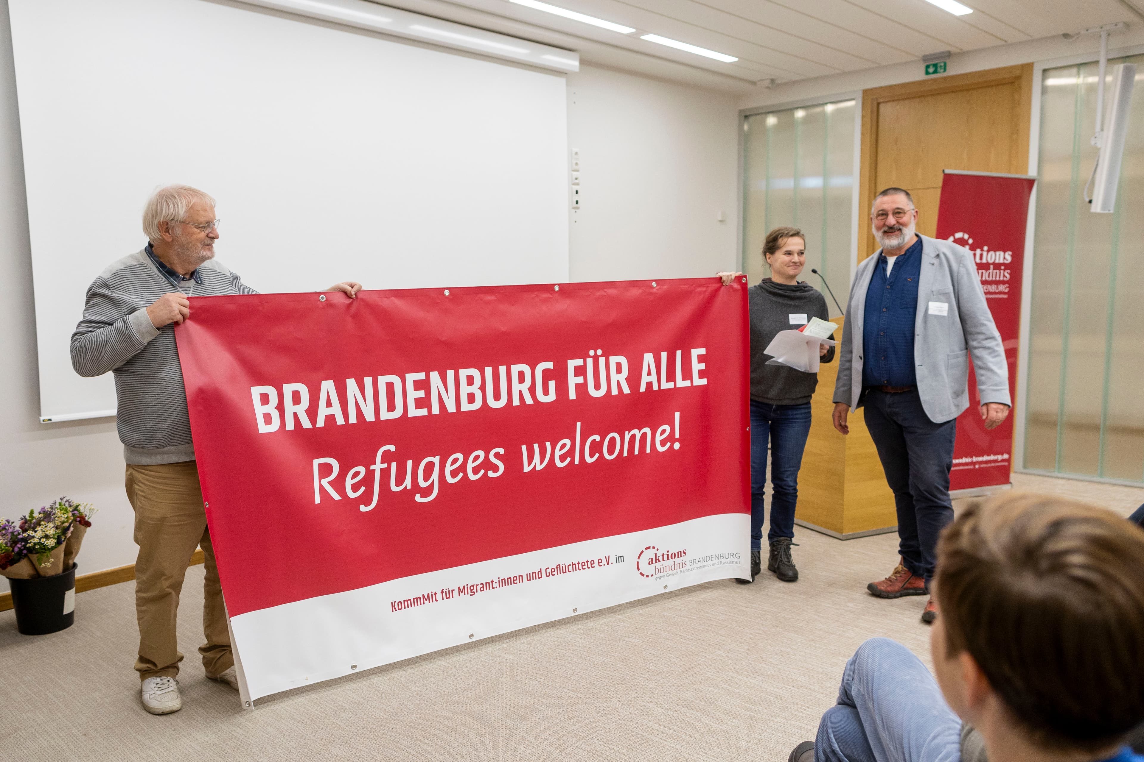 Drei Personen stehen neben einem Banner, auf dem "Brandenburg für alle, Refugees welcome! steht"
